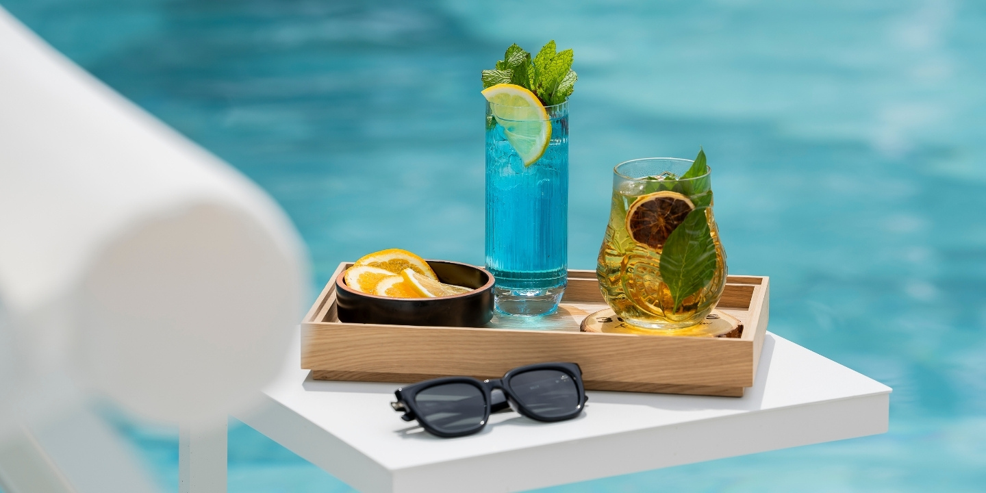 Two colorful drinks, one blue with lemon and mint, the other amber with citrus and basil, sit on a tray by a pool, with sunglasses beside them.