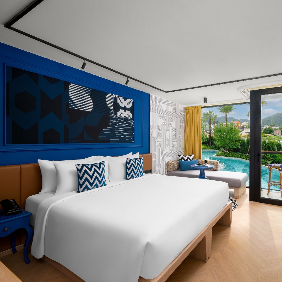 Modern hotel room featuring a king bed, blue accents, patterned cushions, and a cozy seating area overlooking a pool and garden.