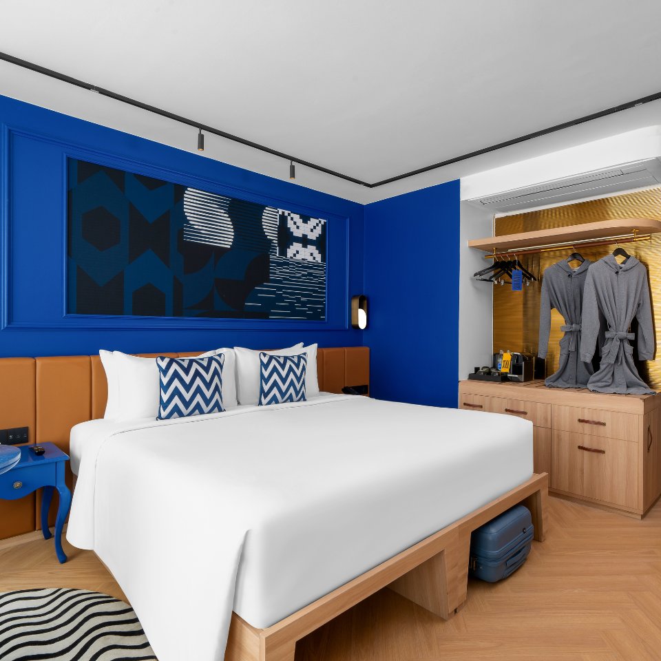 Modern hotel room with a plush white bed, blue and white chevron pillows, deep blue walls, and abstract art. Cozy robes hang beside a stylish open wardrobe.