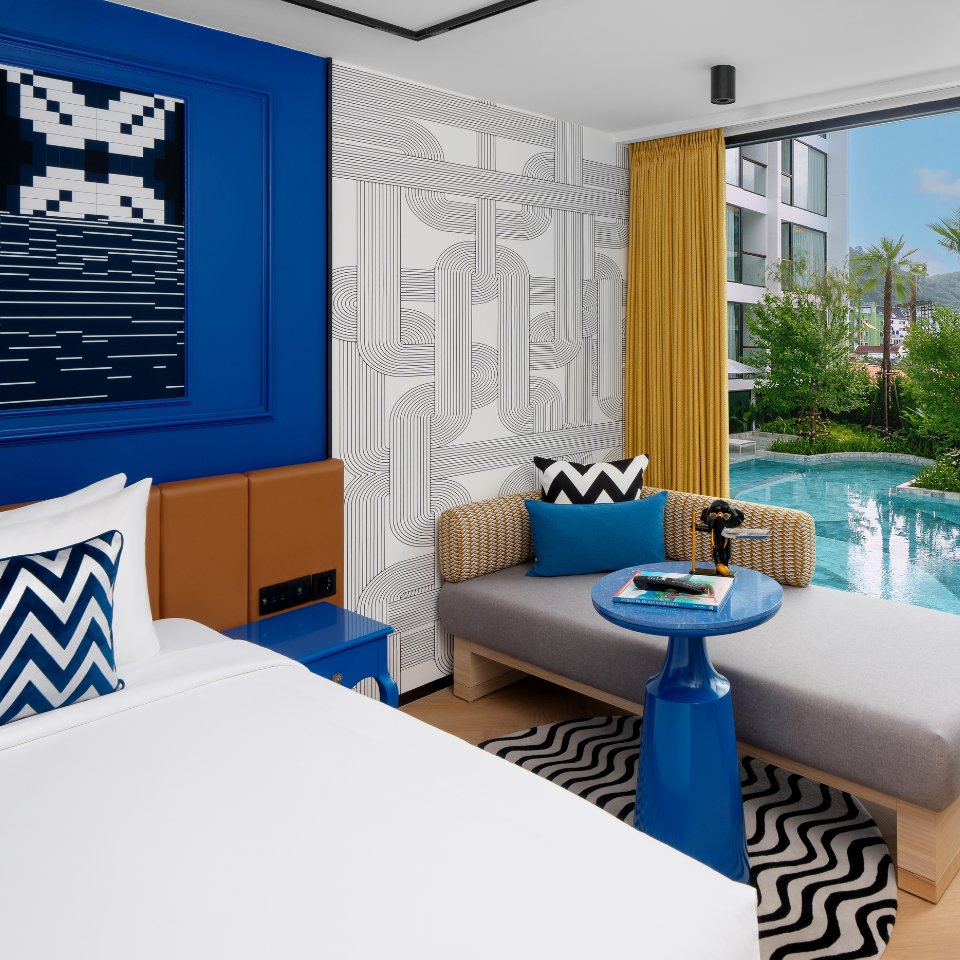 Modern hotel room with bold geometric decor in blue, white, and brown tones. Features a poolside view, lounge area, and zigzag patterns. Bright and inviting.