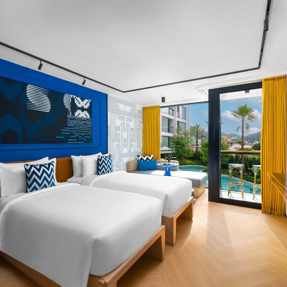 Modern hotel room with two white beds, blue accents, and patterned pillows. Bright yellow curtains frame a view of a balcony, pool, and palm trees.