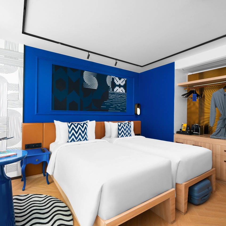 Modern hotel room with two neatly made single beds with white linens, blue and white chevron pillows. Bold blue and orange accents create a vibrant feel. There's a geometric wall art piece, a bedside table, open wardrobe with a hanging robe, a small coffee machine, and a suitcase below, conveying a cozy yet stylish atmosphere.