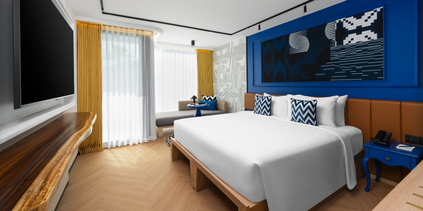 Modern hotel room with a large white bed, blue and white zigzag pillows, vibrant blue artwork, yellow curtains, and a cozy window seat. Bright, inviting atmosphere.