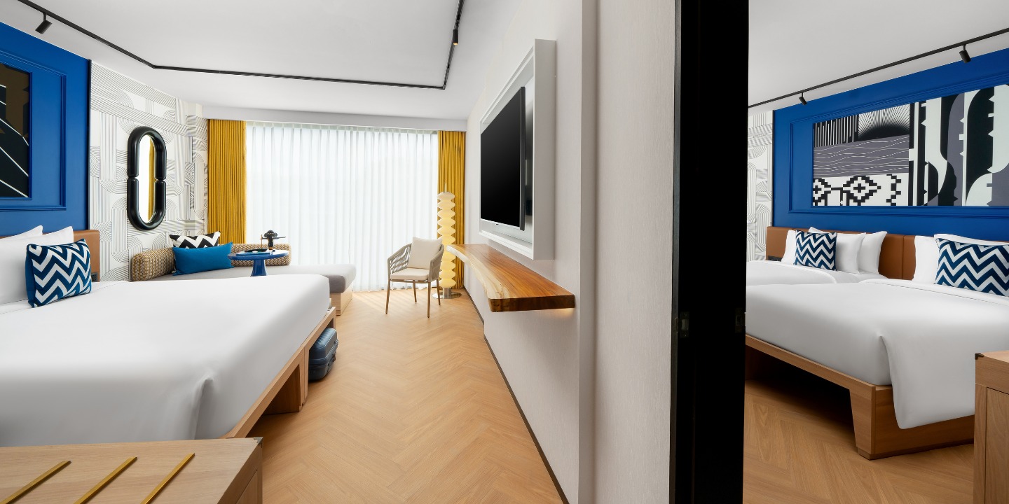 Bright hotel room featuring two beds, modern decor, a flat-screen TV, window with sheer curtains, and stylish seating.