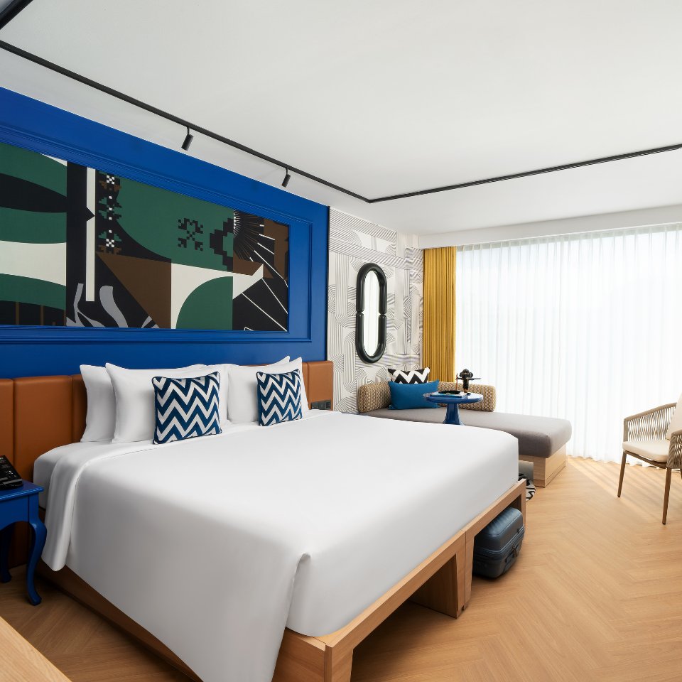 A modern hotel room featuring two beds, vibrant blue accents, large windows with yellow curtains, and stylish decor.