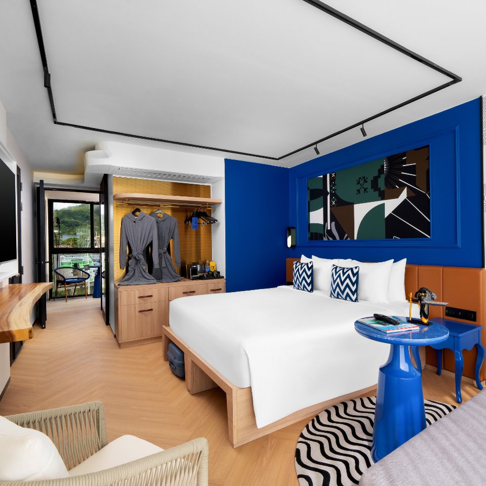 Modern hotel room with a cozy bed, abstract art, and blue accents. Open wardrobe with robes, a balcony view, and a relaxed, inviting atmosphere.