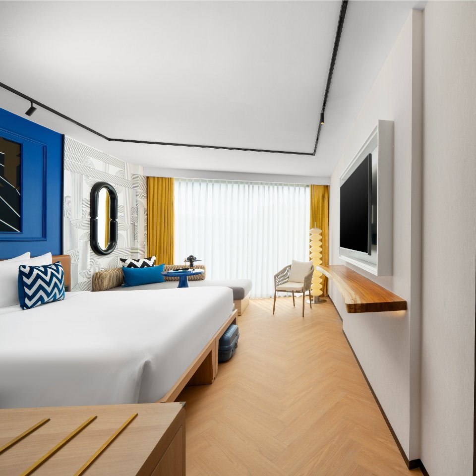 Bright hotel room featuring two beds, modern decor, a flat-screen TV, window with sheer curtains, and stylish seating.