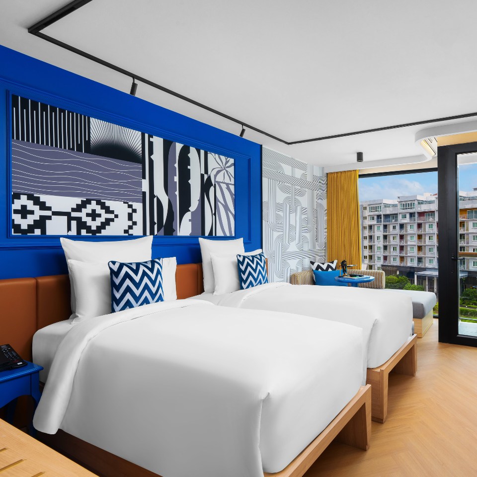 Modern hotel room with two neatly made twin beds featuring white linens and blue zigzag pillows. A vibrant blue accent wall displays abstract art. Large windows with yellow curtains overlook a cityscape, creating a bright, inviting atmosphere.