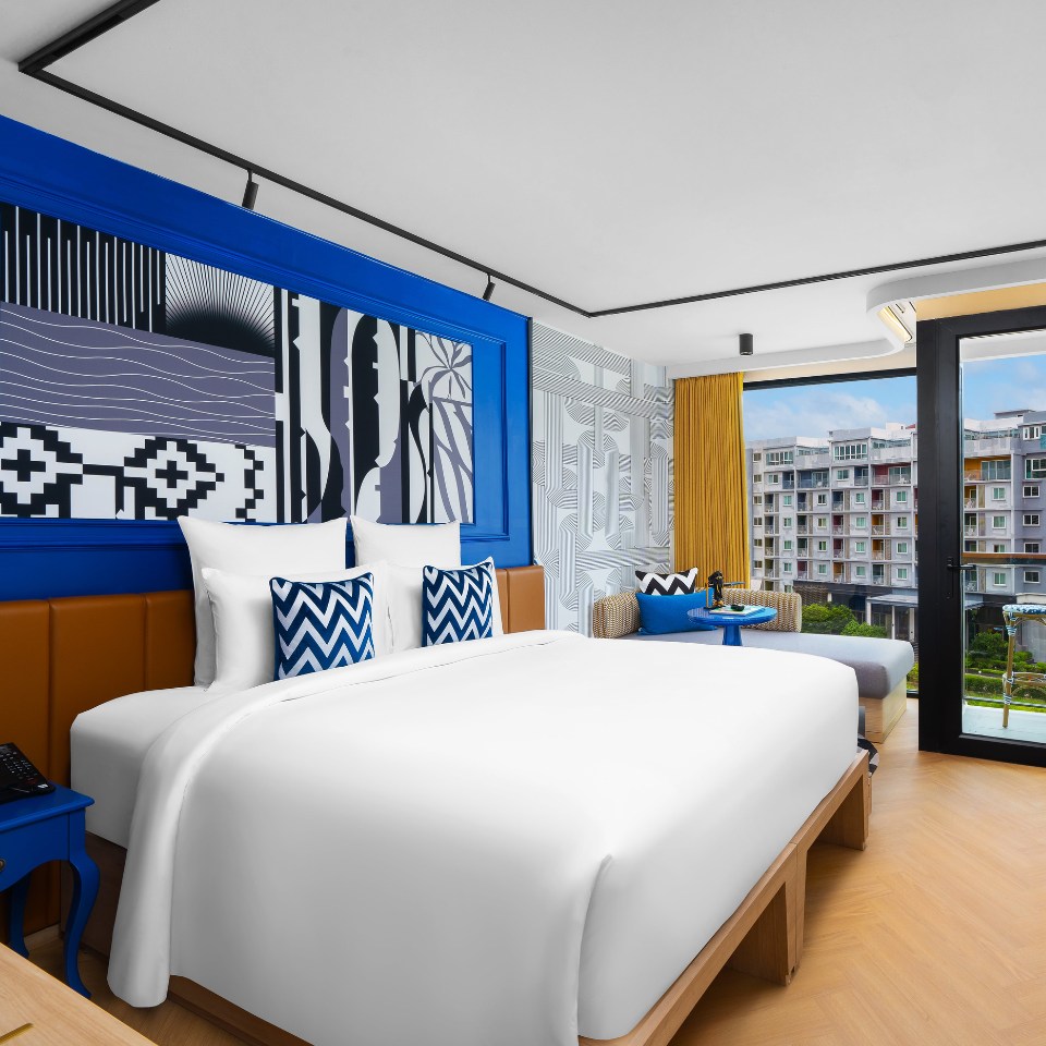 Modern hotel room with a large white bed, vibrant blue accents, abstract wall art, chevron pillows, and a view of an urban landscape through a window.