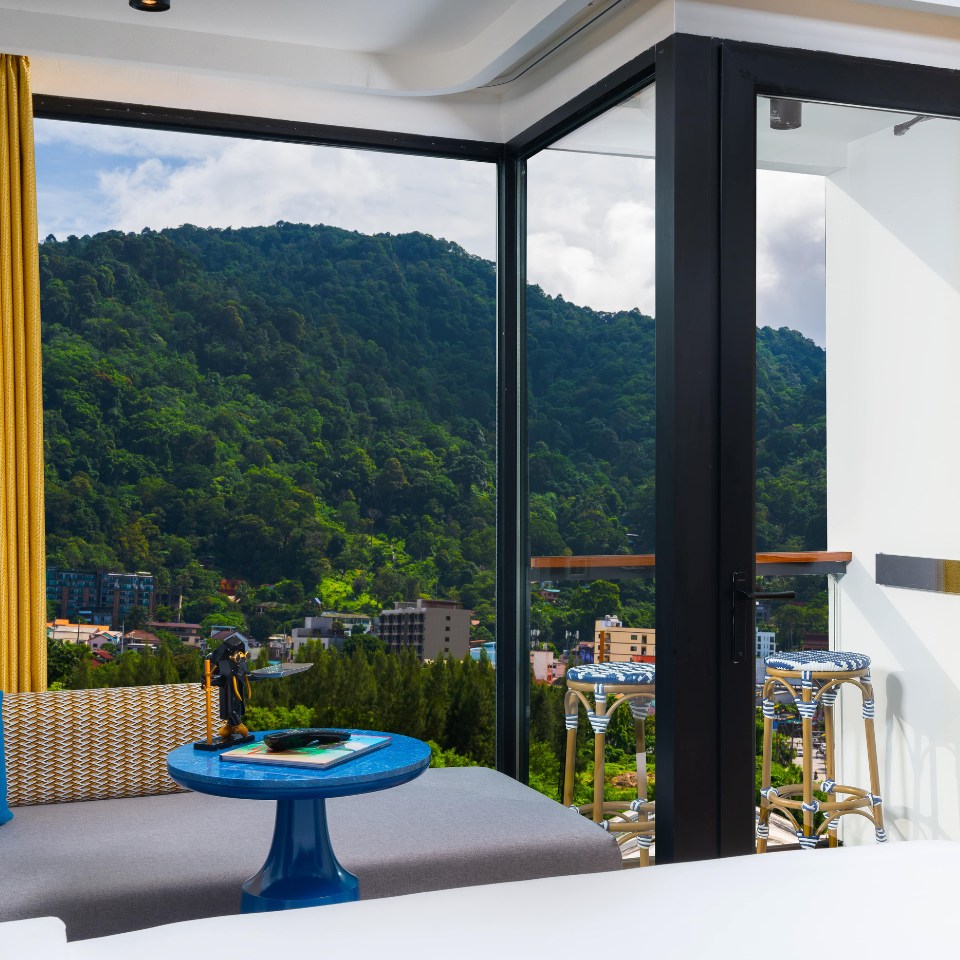 Modern room with a mountain view through floor-to-ceiling windows. There's a cozy seating area, a blue table, and a warm, inviting atmosphere.