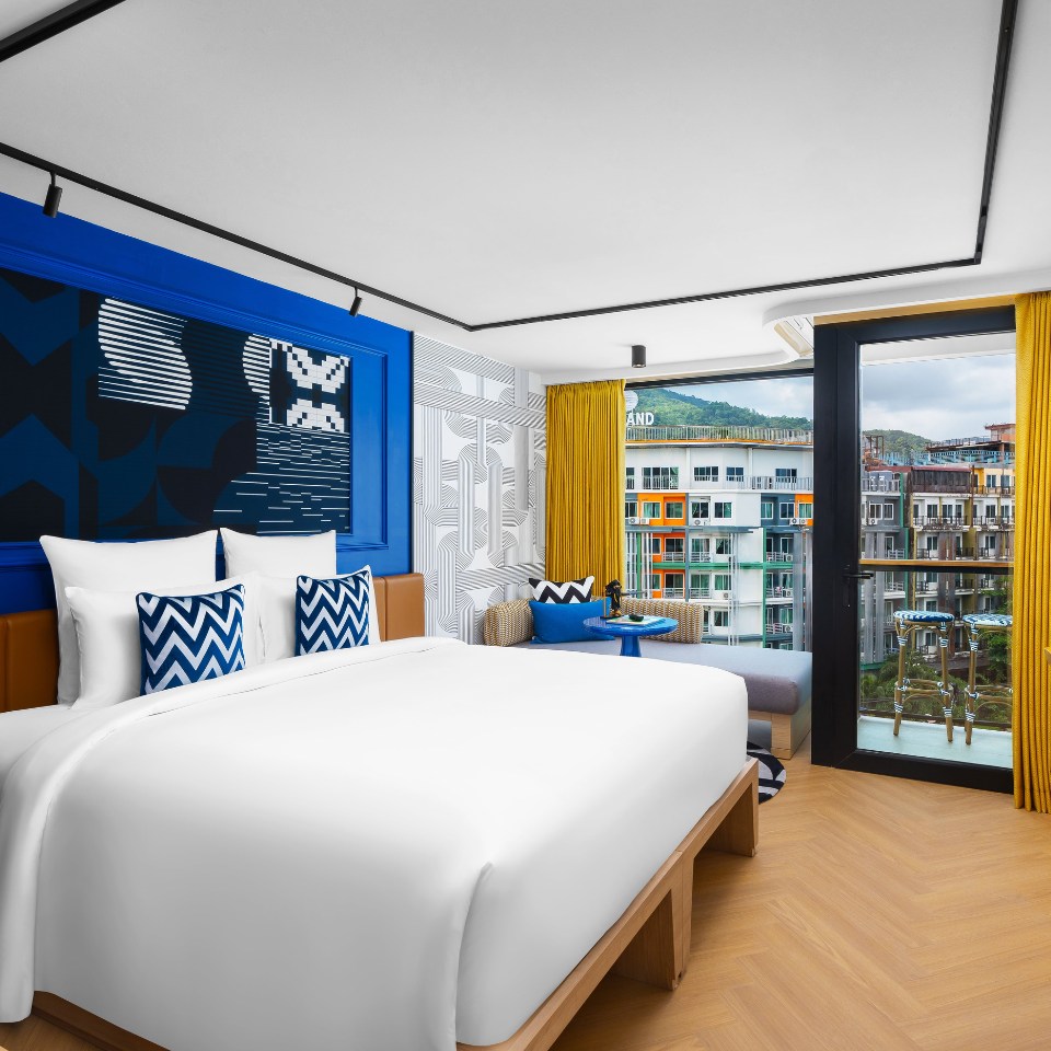 Bright hotel room with a large white bed, blue and white decorative pillows, abstract wall art, and yellow curtains framing a cityscape view.