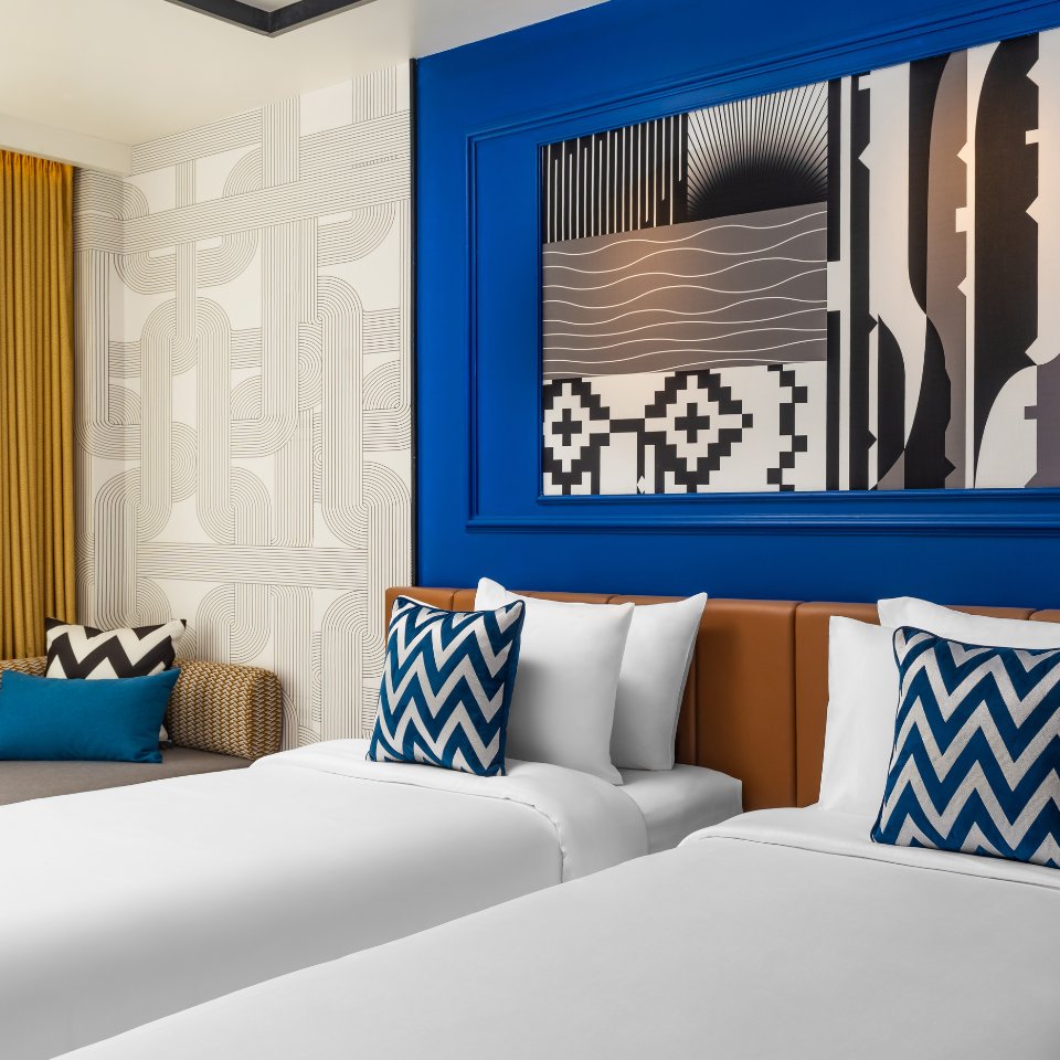 Modern hotel room with two white beds featuring blue zigzag pillows. Bold artwork with geometric patterns on vibrant blue wall. Cozy and stylish ambiance.