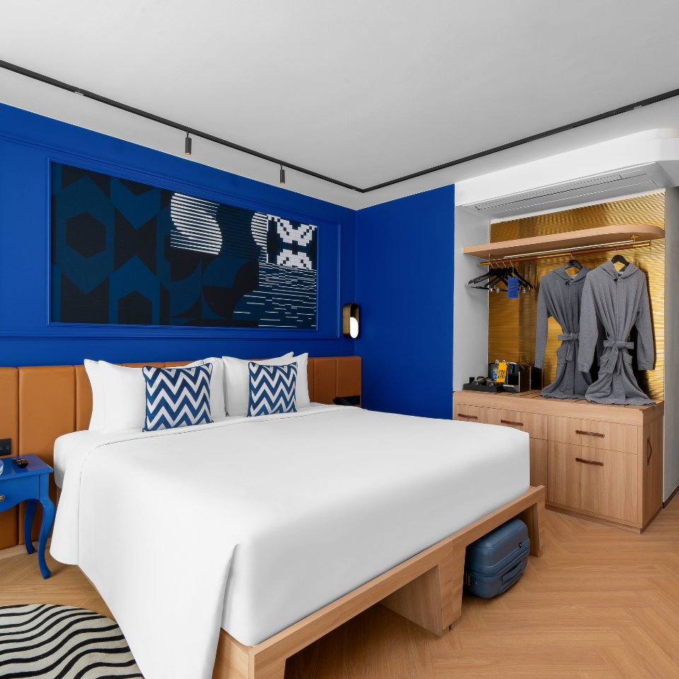 Modern hotel room with a plush white bed, blue and white chevron pillows, deep blue walls, and abstract art. Cozy robes hang beside a stylish open wardrobe.