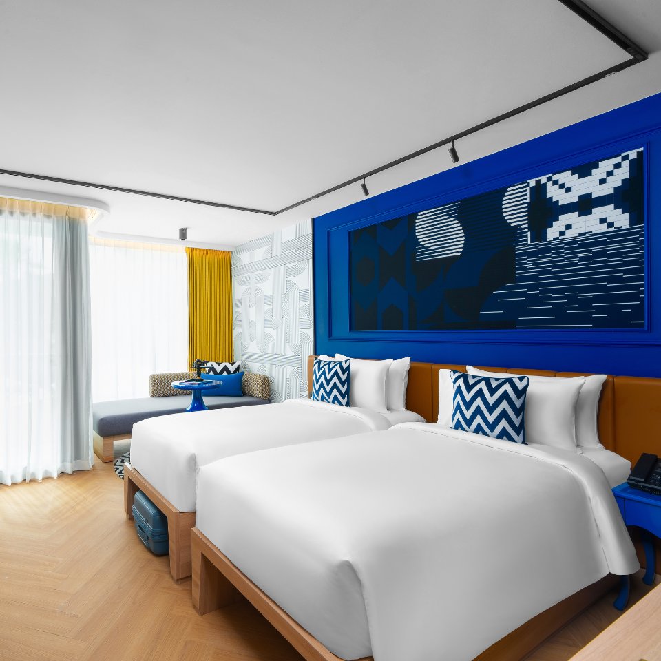 Modern hotel room with two white beds, blue and white patterned pillows, bright artwork above, and a window with sheer yellow and white curtains.
