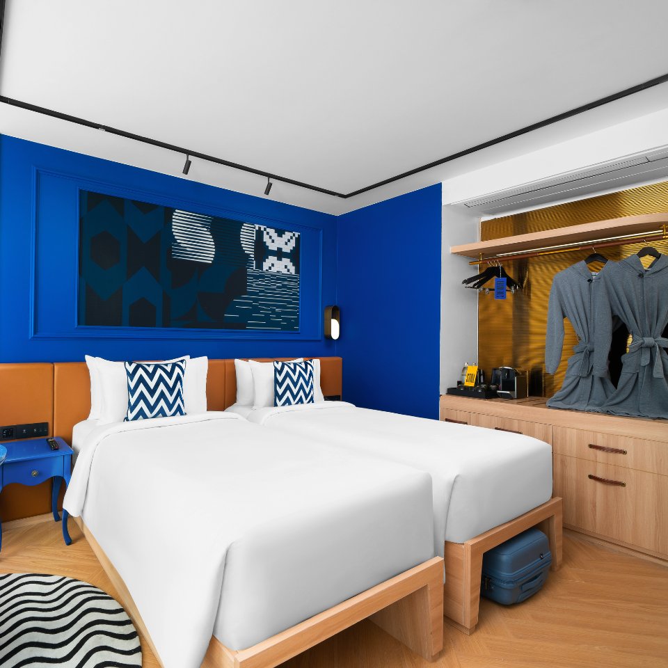 Modern hotel room with two white twin beds, vibrant blue walls, and abstract wall art. Features patterned pillows, robes, and a small coffee station.