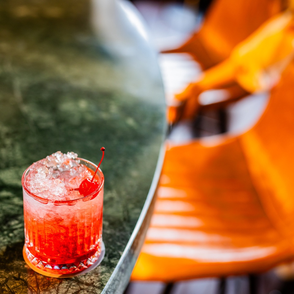 A close up of a red drink with crushed ice on a marble surface. There is a cocktail cherry on top.