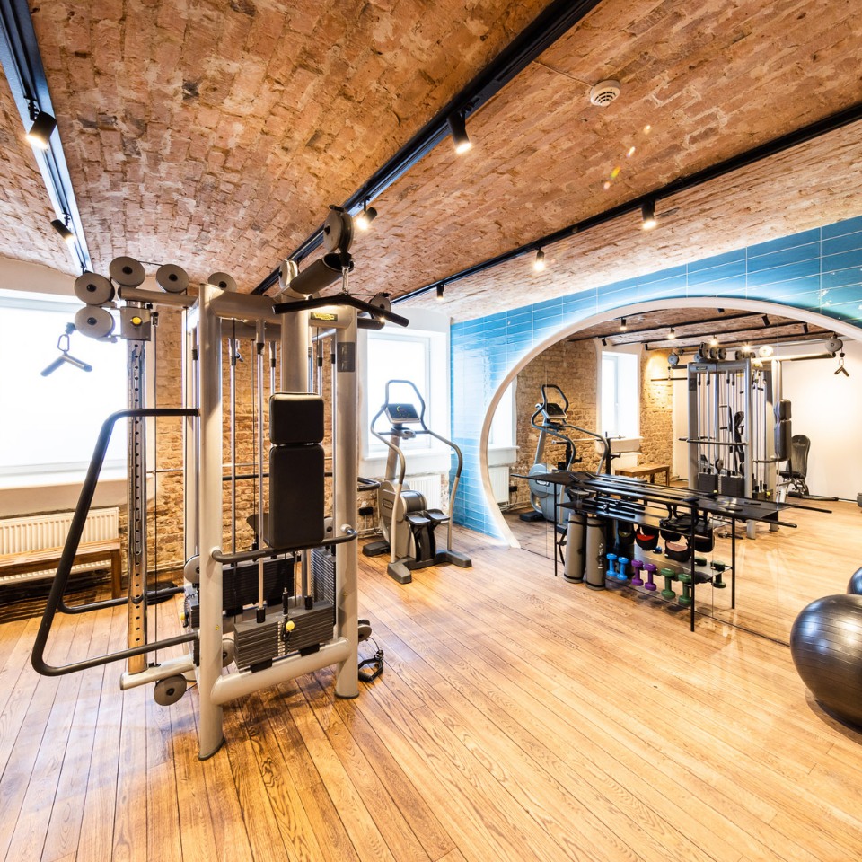A modern gym with blue tiles, featuring exercise equipment, weights, and stability balls visibile in a circular mirror.