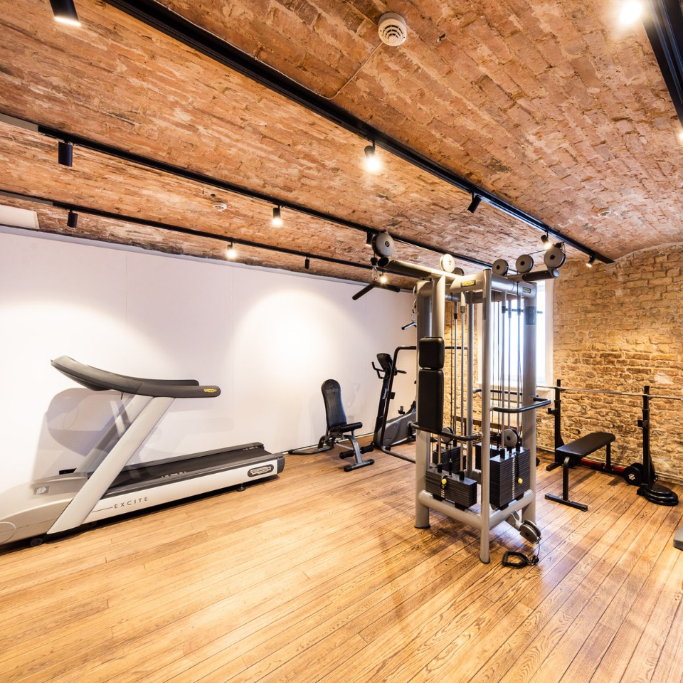 A modern gym interior featuring various exercise machines, weights, and a brick wall, illuminated by track lighting.