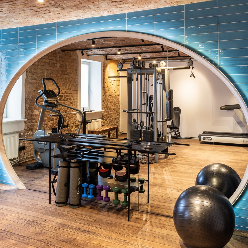 A modern gym with blue tiles, featuring exercise equipment, weights, and stability balls visibile in a circular mirror.