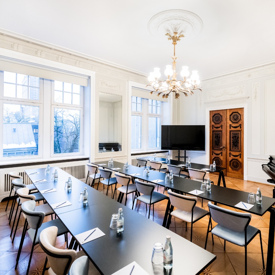 A stylish conference room featuring long tables, modern chairs, a TV, and elegant decor, illuminated by a chandelier and natural light.
