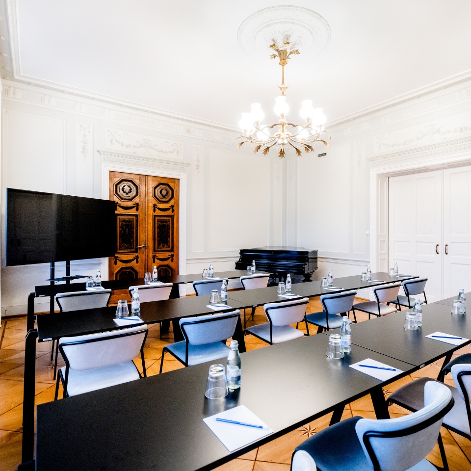 A stylish conference room featuring long tables, modern chairs, a TV, and elegant decor, illuminated by a chandelier and natural light.