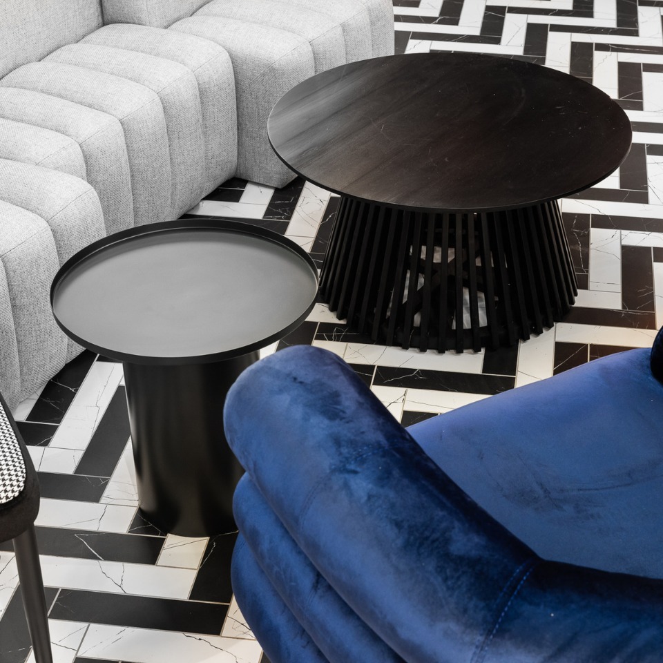 A modern living space featuring a gray sofa, black circular tables, and a blue velvet chair, set on a patterned black-and-white floor.
