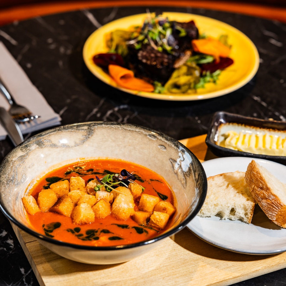 A bowl of vibrant tomato soup topped with croutons and microgreens, accompanied by bread and butter, with a plated dish in the background.