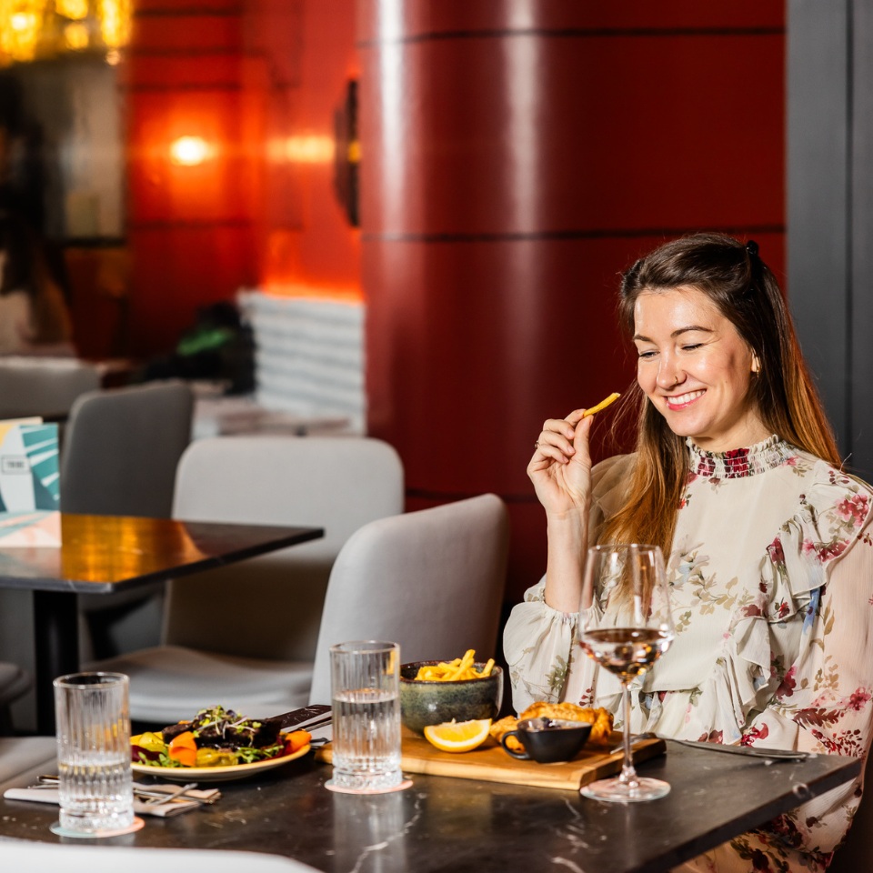 A woman holds a fry, seated at a table set with a salad, drinks, and a platter of food, in a stylish restaurant.