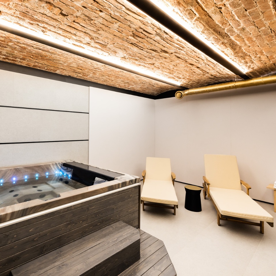 A modern spa room featuring a wooden hot tub, two loungers, and a rustic brick ceiling with stylish lighting.