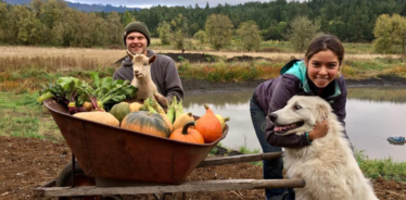 Man and a woman with dog posting for the camera next to a wheelbarrow full of pumpkins and gourds